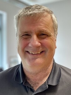 Herr Mackenzie mit grauem Haar, in Polohemd lächelnd.